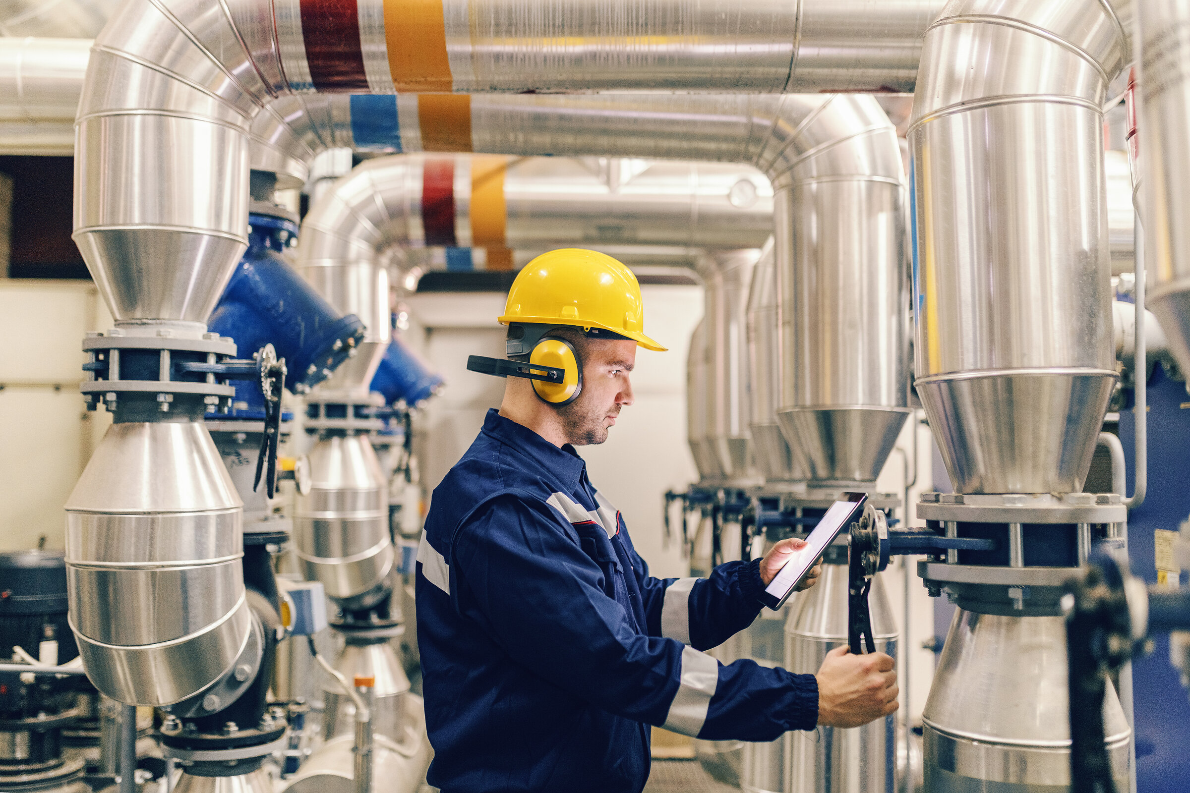 Profile of young Caucasian worker tightening the valve and using tablet while standing in heating plant.